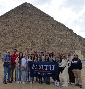 Students and Faculty hold up a Utica University banner at the Great Pyramid during a January 2023 visit to Egypt.